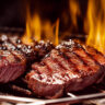 Grilled meat steak on stainless grill depot with flames on dark background. Food and cuisine concept.